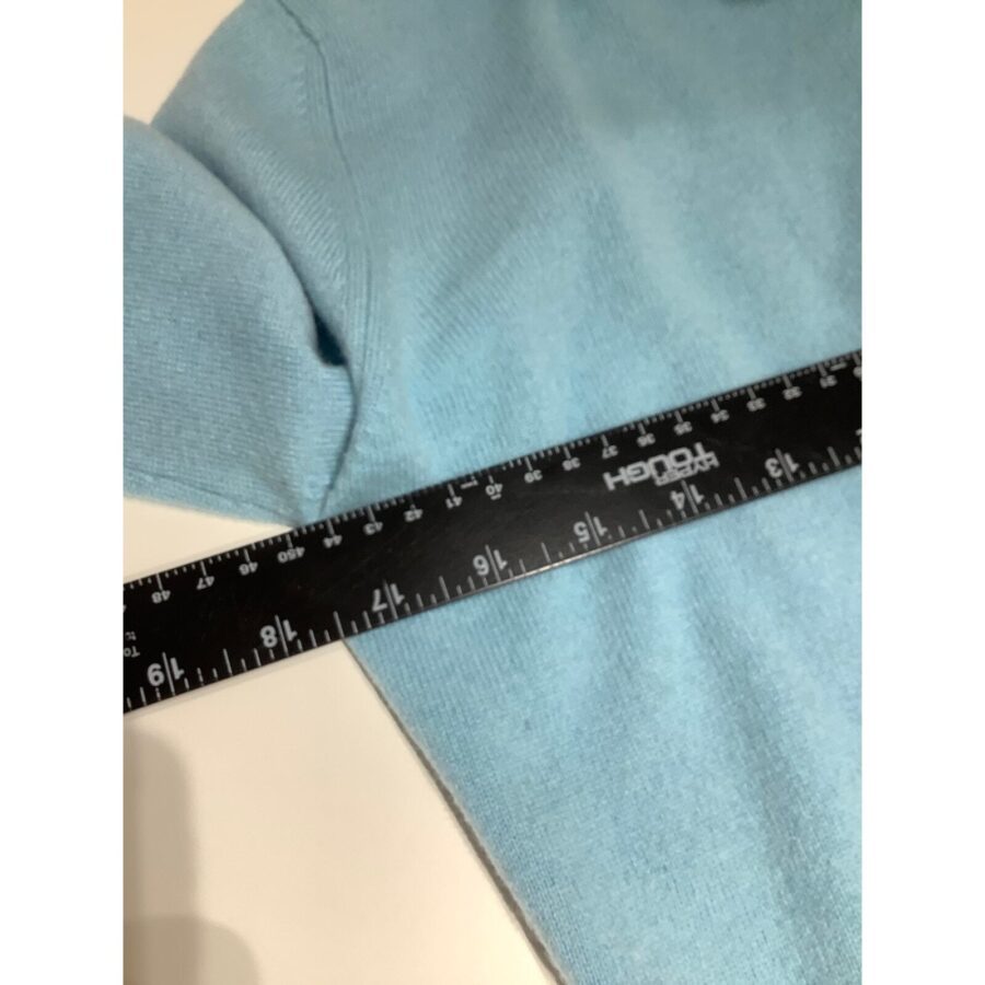 A close-up image showing a black ruler measuring a light blue garment, likely a sweater, against a white background.