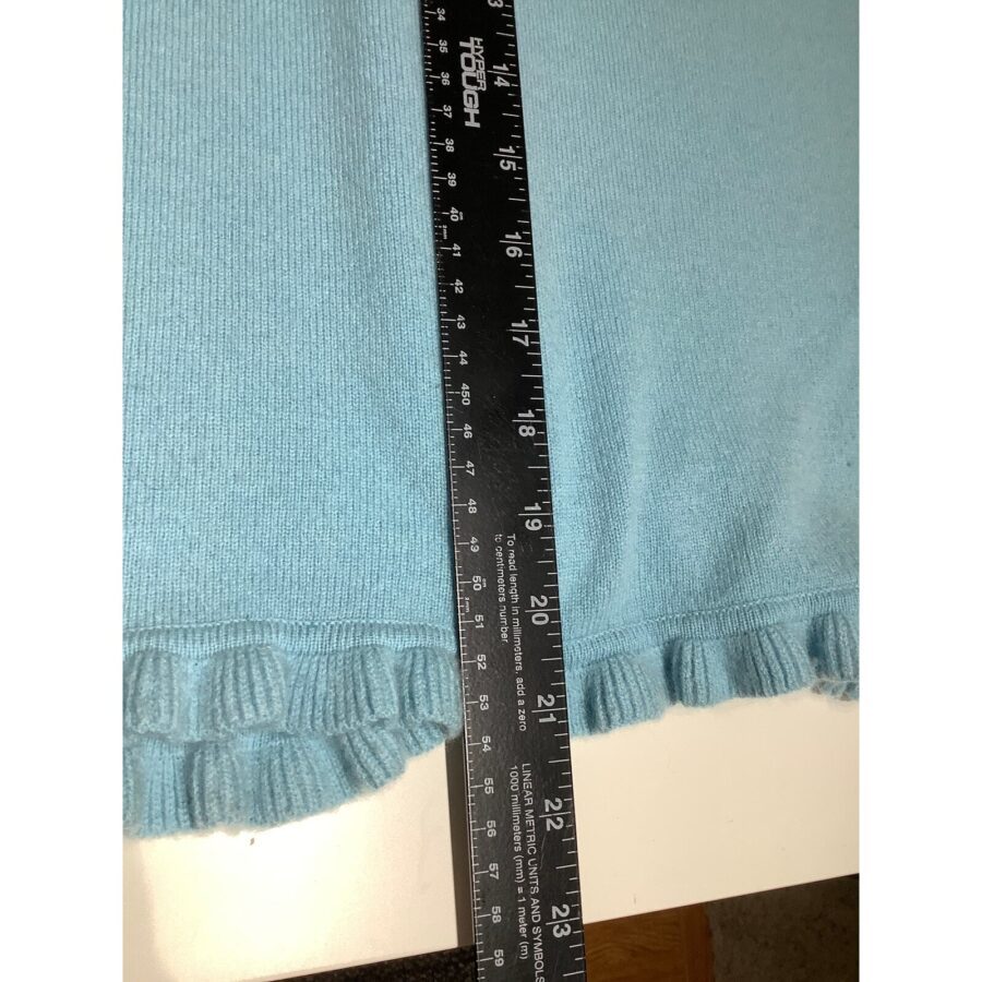 A light blue knit garment with ruffled edges is measured by a black ruler, displaying inches and centimeters, placed on a white surface.