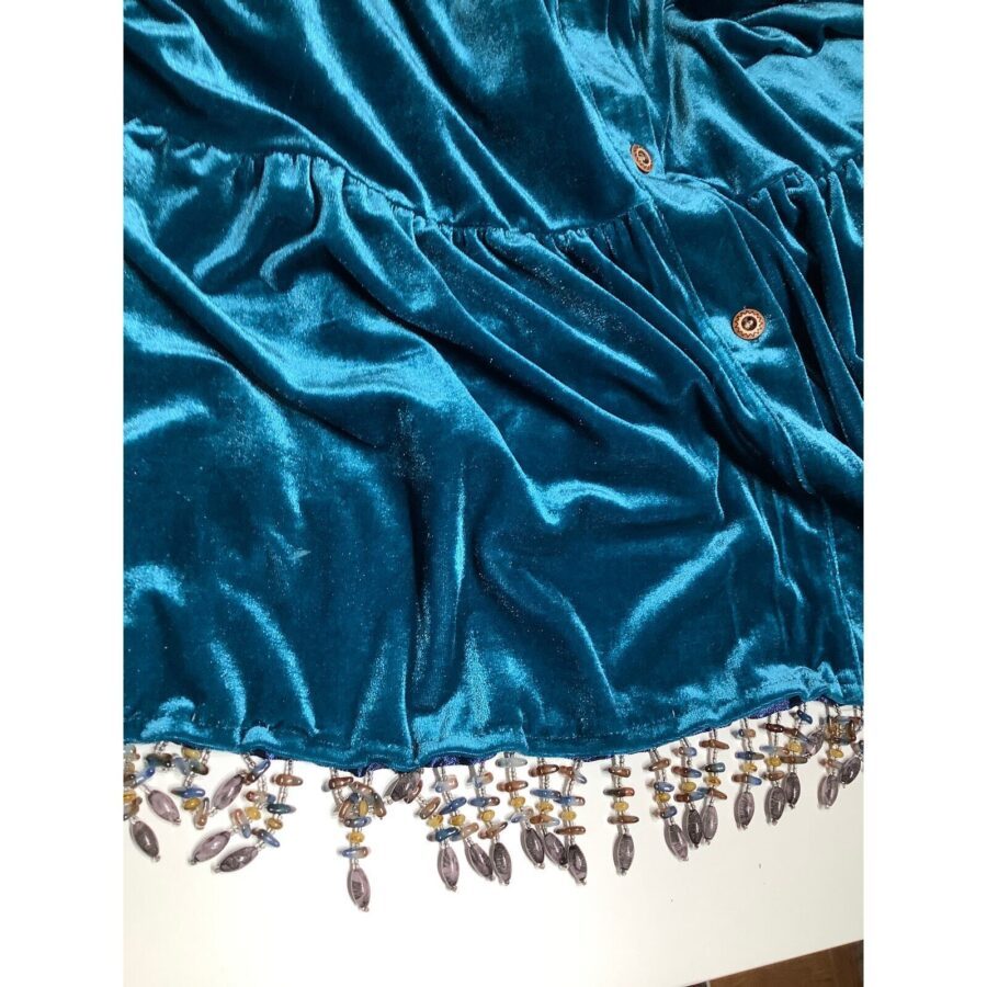 The image shows a close-up of a teal silk fabric with buttons and intricate dangling beadwork on its edge, laid out flat.